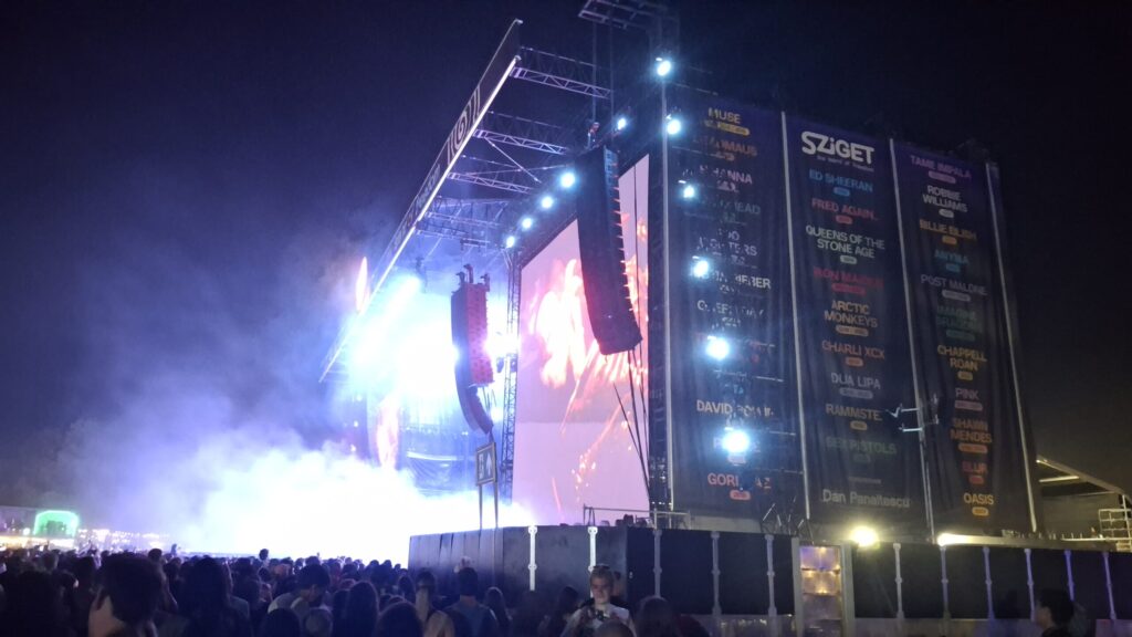 Nachtaufnahme einer großen Festivalbühne beim Sziget Festival. Scheinwerfer und LED-Leinwände beleuchten die Bühne, während sich davor eine dichte Menschenmenge versammelt. An der seitlichen Bühnenkonstruktion hängen großformatige Banner mit dem Sziget-Logo und Namen verschiedener Künstler.